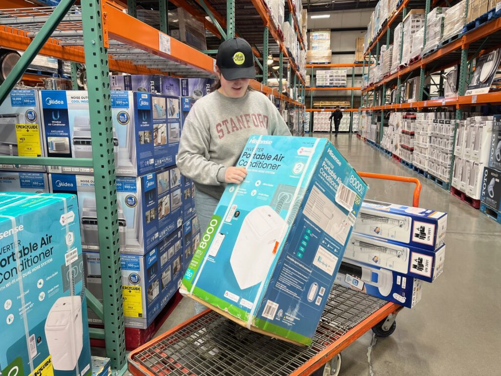 A Meals on Wheels People intern in a Stanford sweatshirt and black cap loads a boxed portable air conditioner onto a flat cart in a warehouse store aisle filled with appliances.