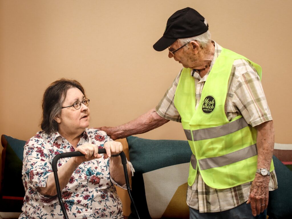 Older man, wearing a yellow safety vest and hat, gently touches the shoulder of a seated woman with a walker, both looking at each other with concern.