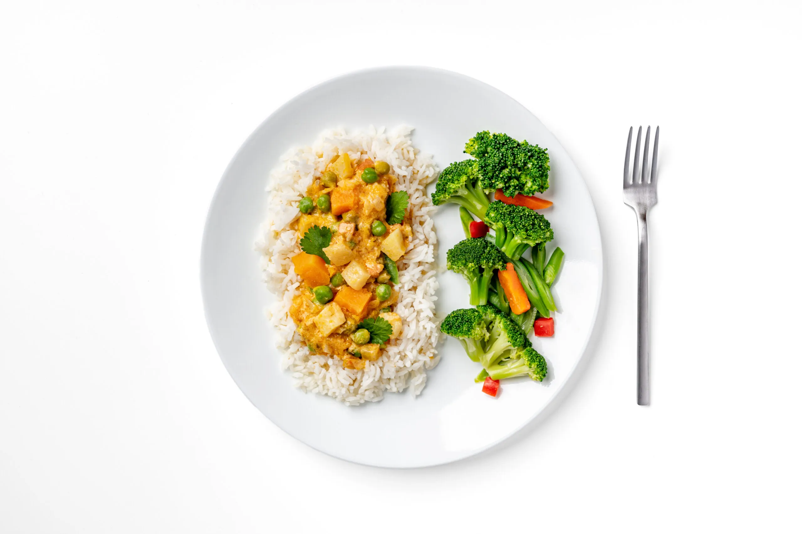 A white plate with white rice topped with butternut squash and chickpea curry and vegetables, served with a side of steamed broccoli, green beans, and carrots. A fork is placed to the right of the plate on a white background.