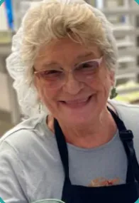 Deanna Plewik, short gray hair and glasses, smiles warmly. She's wearing a blue shirt and black apron, standing in a bright kitchen, exuding a joyful, welcoming vibe.