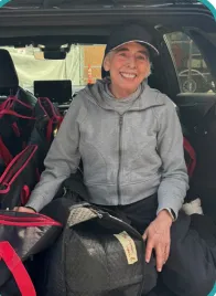 Debi Stedron in a gray jacket and cap smiles while sitting in a car trunk surrounded by Meals 4 Kids delivery bags. The scene is cheerful and casual, conveying warmth.