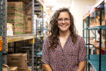 A woman with curly brown hair and glasses, wearing a floral shirt, stands smiling in a warehouse aisle with shelves of boxes and supplies in the background.