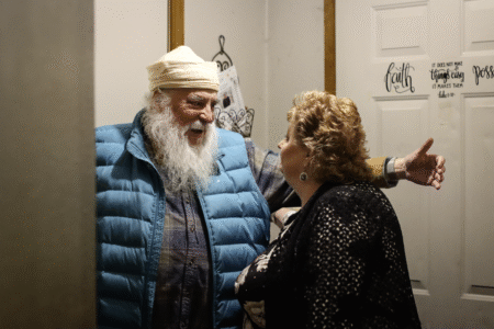 An older man in a blue vest and white cap joyfully embraces a woman in a black outfit. They stand in a warmly lit room beside a door with inspirational quotes.