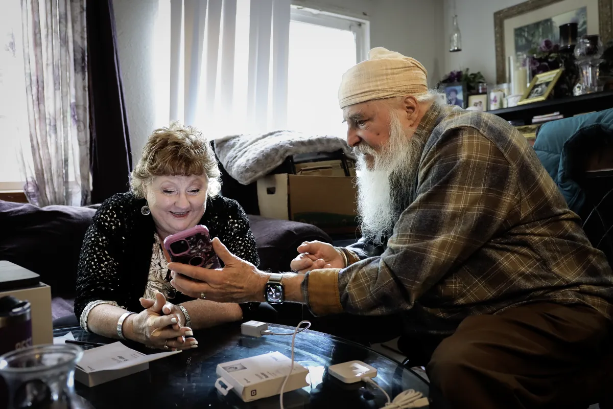 An older man and woman share a joyful moment, smiling at a smartphone the man is holding. They are seated together in a cozy, well-lit room.