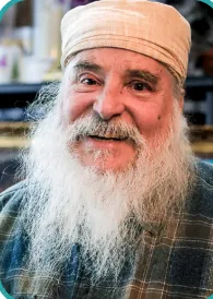 Guruka Singh with a white beard and a gentle smile, wearing a tan turban and plaid shirt. The background is softly blurred.
