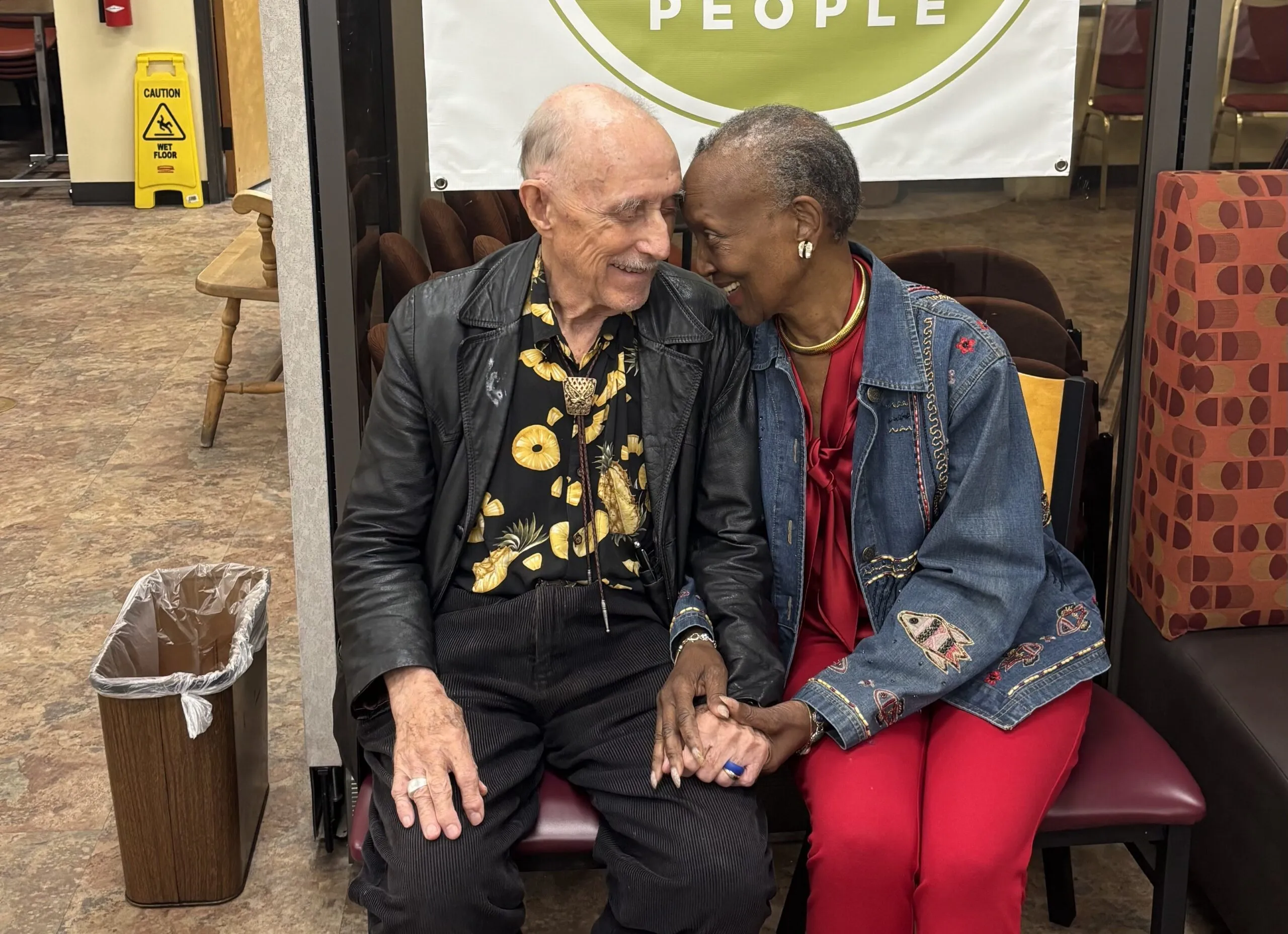 An older couple sits closely on a bench, holding hands and smiling warmly at each other. They are dressed in colorful outfits, conveying love and joy.