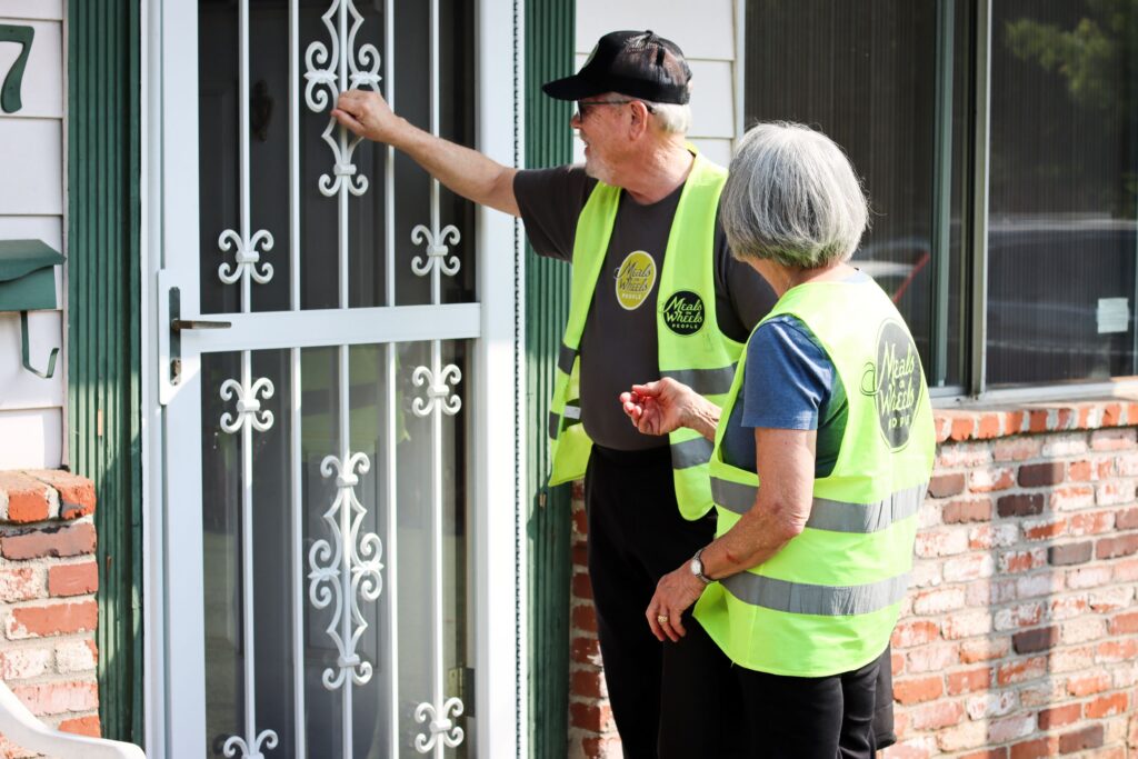 Two Meals on Wheels People volunteers in yellow vests stand at a door, with one person knocking. They appear focused and engaged in community service.
