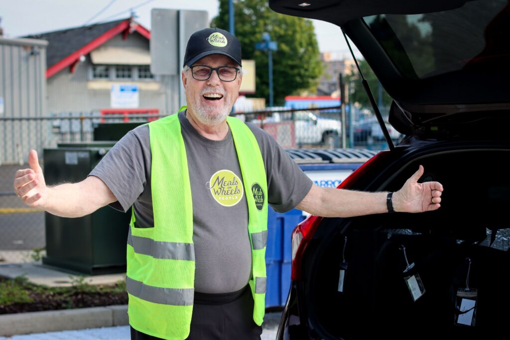 Smiling man in a "Meals on Wheels People" cap and vest, standing next to an open car trunk with meal bags, arms open wide, conveying warmth and enthusiasm.