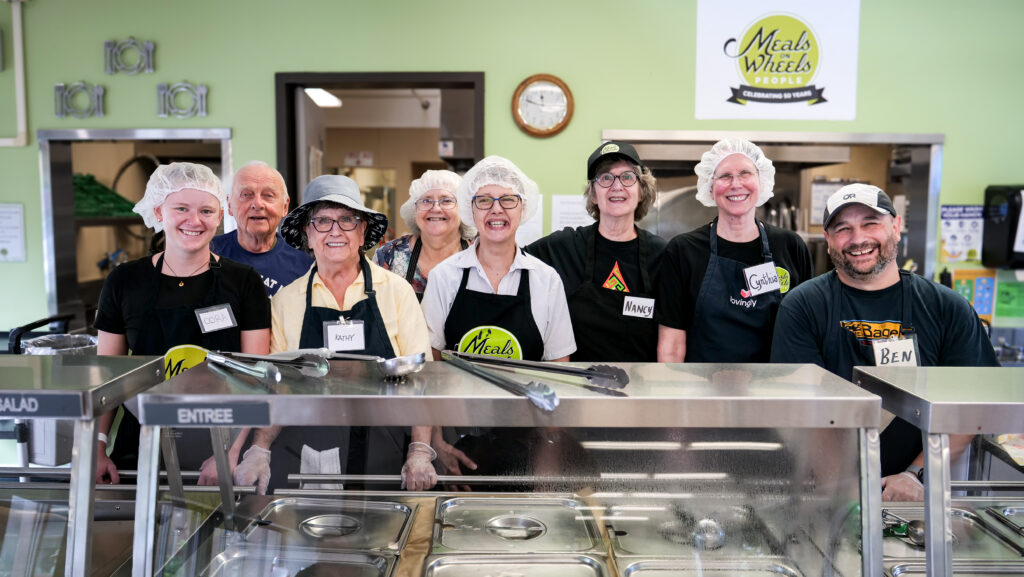 A group of eight smiling volunteers and staff in a kitchen stand behind a food serving counter. They wear aprons and hairnets. A "Meals on Wheels People" sign is visible above.