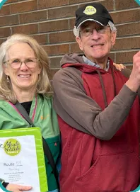 Volunteers Ellen Goldschmidt and Keith Salenger stand together in front of a brick wall at the Meals on Wheels People Southwest Center. Ellen holds a folder with papers inside, and Keith wears a black cap and red hoodie.