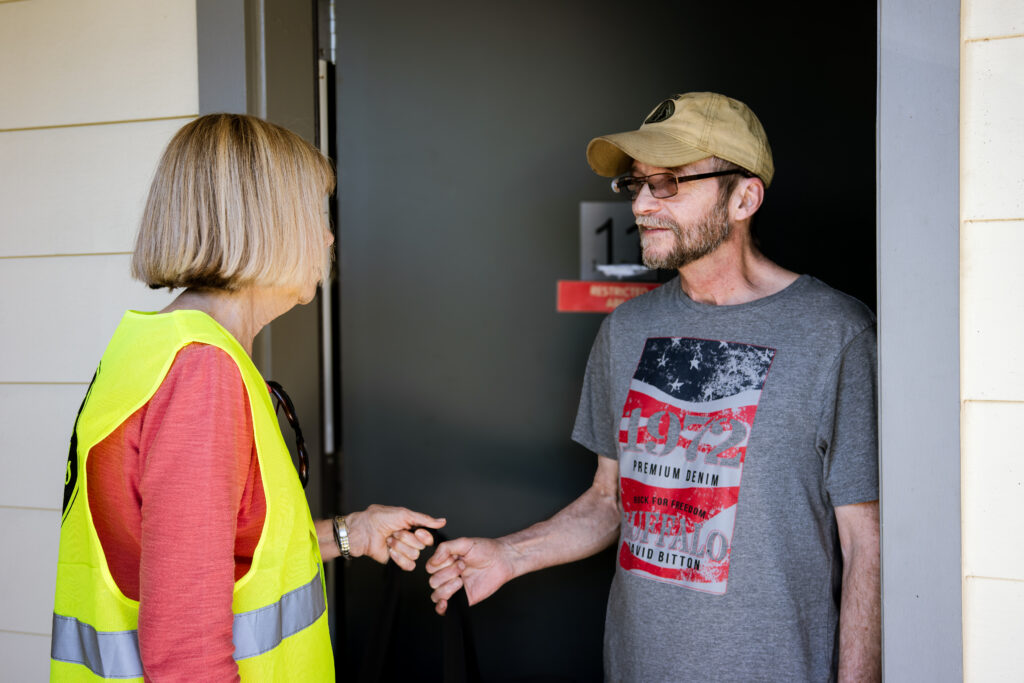 Barbara, a Meals on Wheels People volunteer wearing a yellow safety vest, hands a meal to Larry, a smiling older man standing in the doorway of his home.