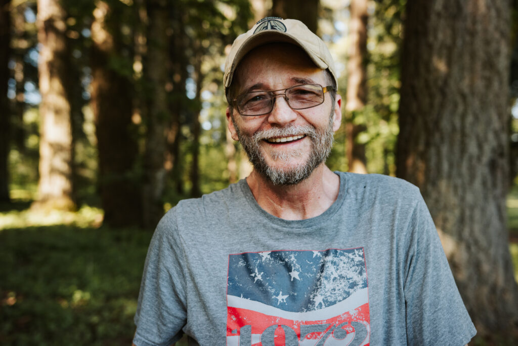 Larry, a smiling older man with a gray beard, glasses, and a beige baseball cap, stands outdoors in a sunlit forest. He’s wearing a gray T-shirt with a weathered American flag design.