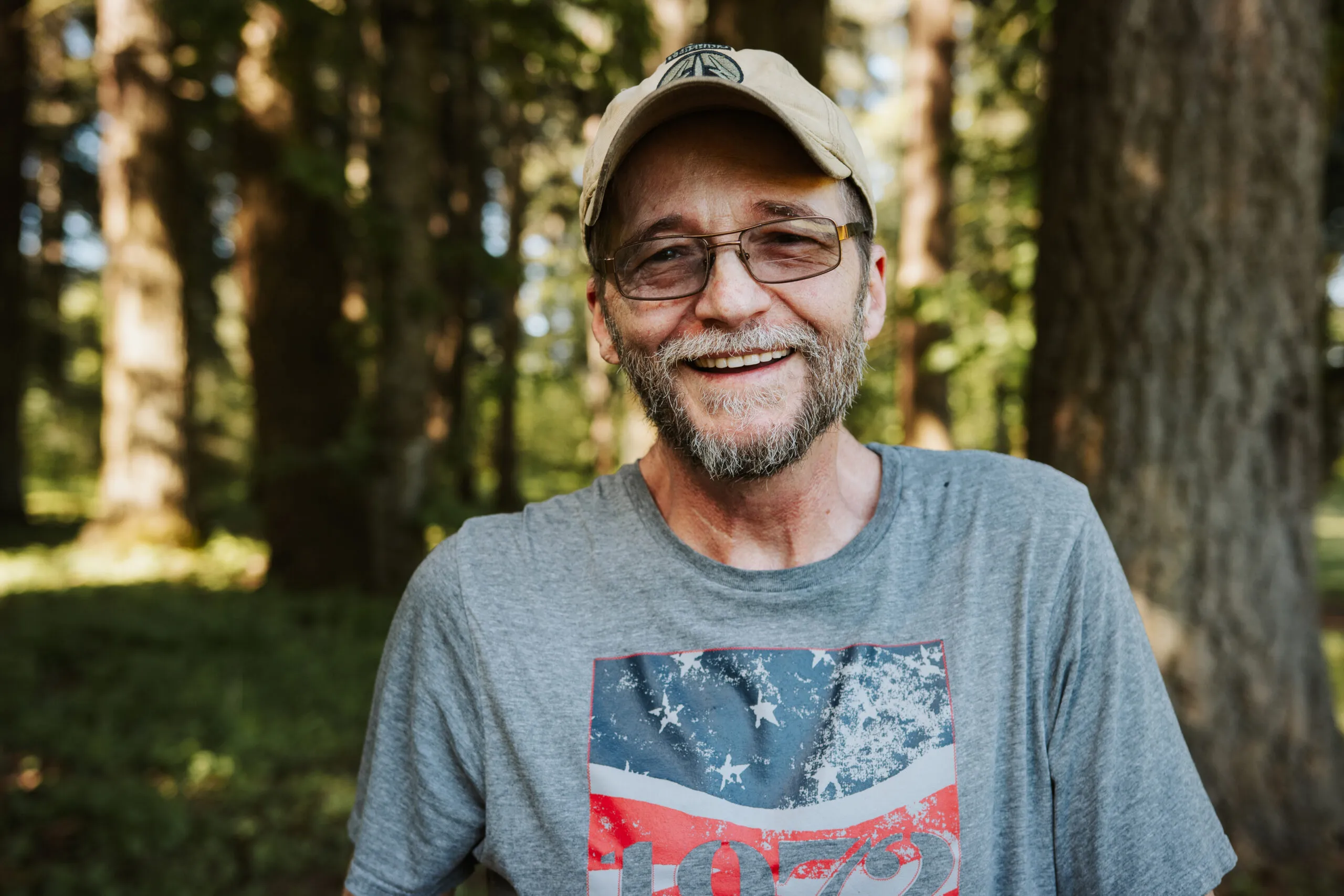 Larry, a smiling older man with a gray beard, glasses, and a beige baseball cap, stands outdoors in a sunlit forest. He’s wearing a gray T-shirt with a weathered American flag design.
