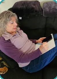 Lauri Howe writes notes on her Friendly Chats calls while on a dark couch, wearing a purple top and jeans, using a smartphone. Her expression is focused, creating a calm atmosphere.