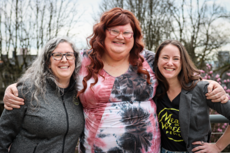 Two Meals on Wheels People staff and Leah stand closely together, smiling warmly with arms around each other. The background features bare trees, hinting at early spring.