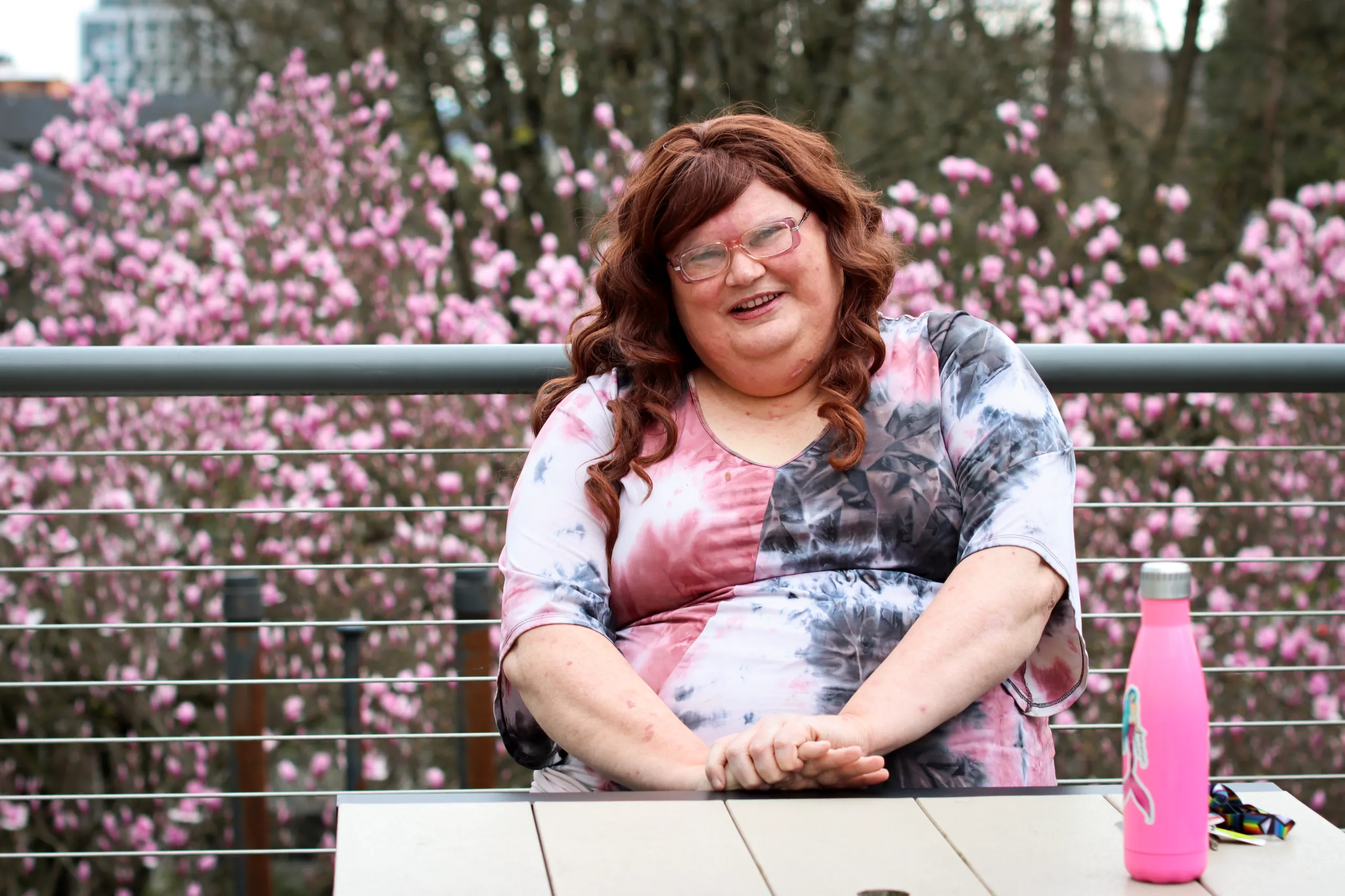Leah, smiling, sits at a table outside, with blooming pink flowers in the background. A pink water bottle is on the table.