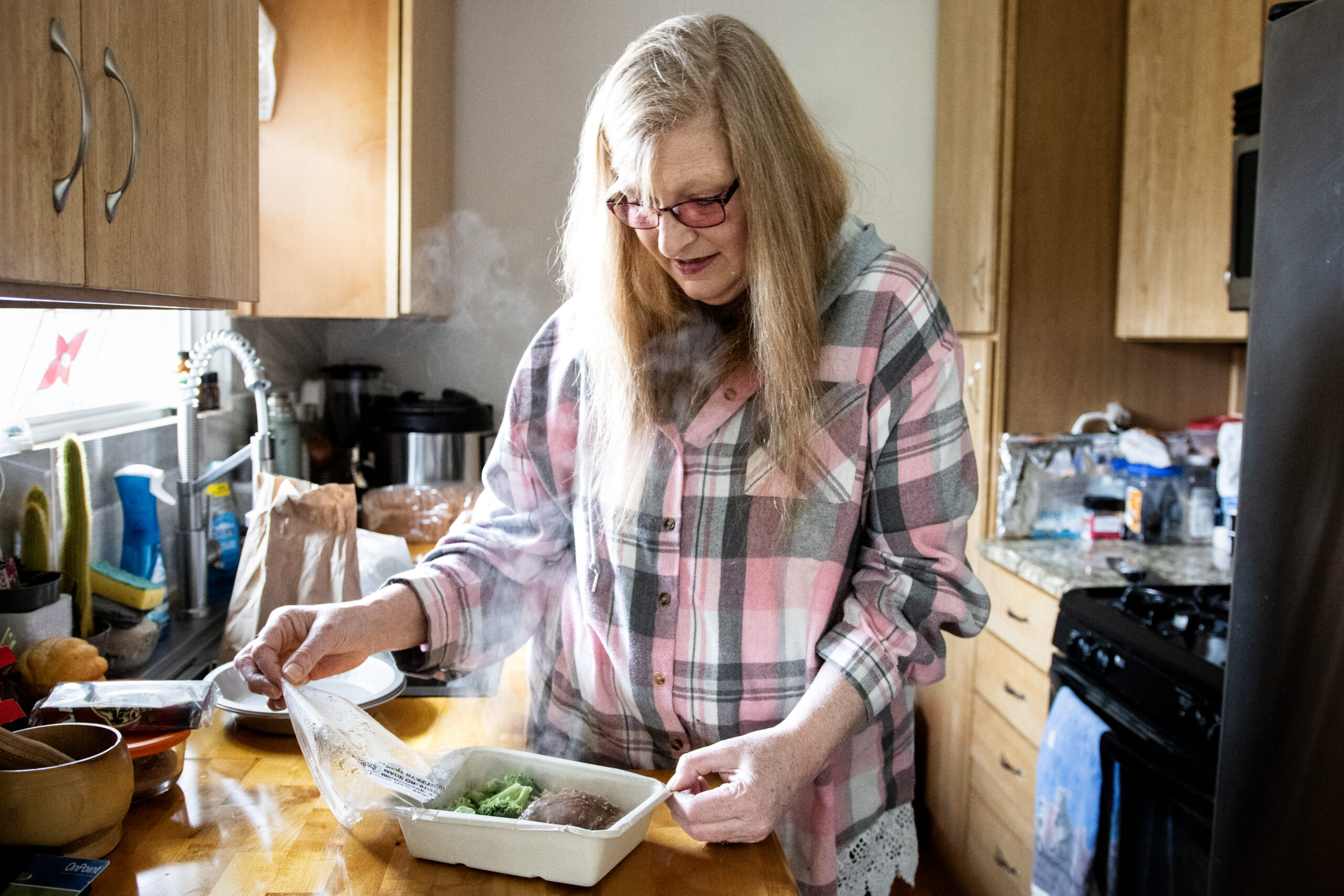 A woman in a plaid shirt stands in a kitchen, peeling back the lid of a steaming meal tray with broccoli and beef. She appears focused and content.