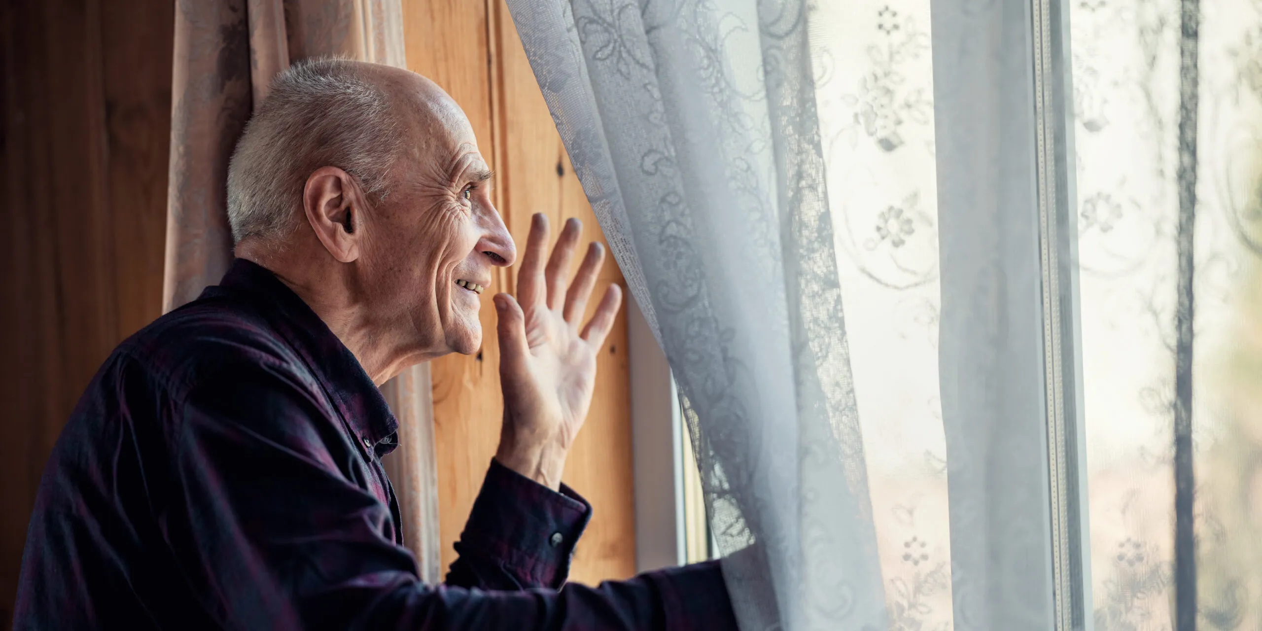 An older man smiles and waves while looking out a window with sheer, patterned curtains in a warmly lit room.