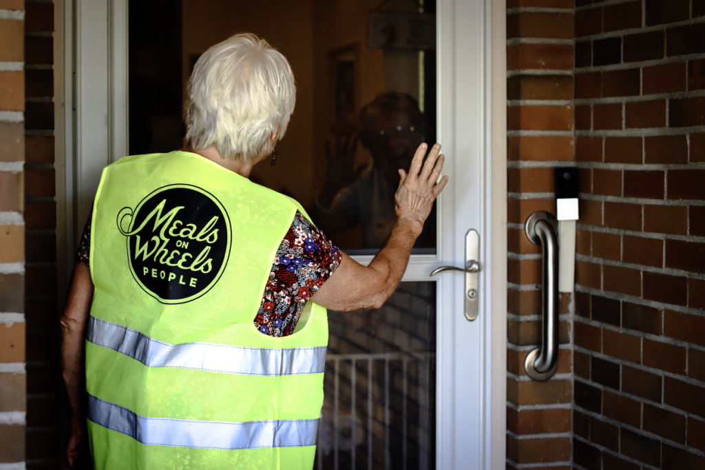 A Meals on Wheels People volunteer wearing a high-visibility vest stands at a door, reaching out with one hand as if to knock or greet someone inside. The vest has the logo "Meals on Wheels People."