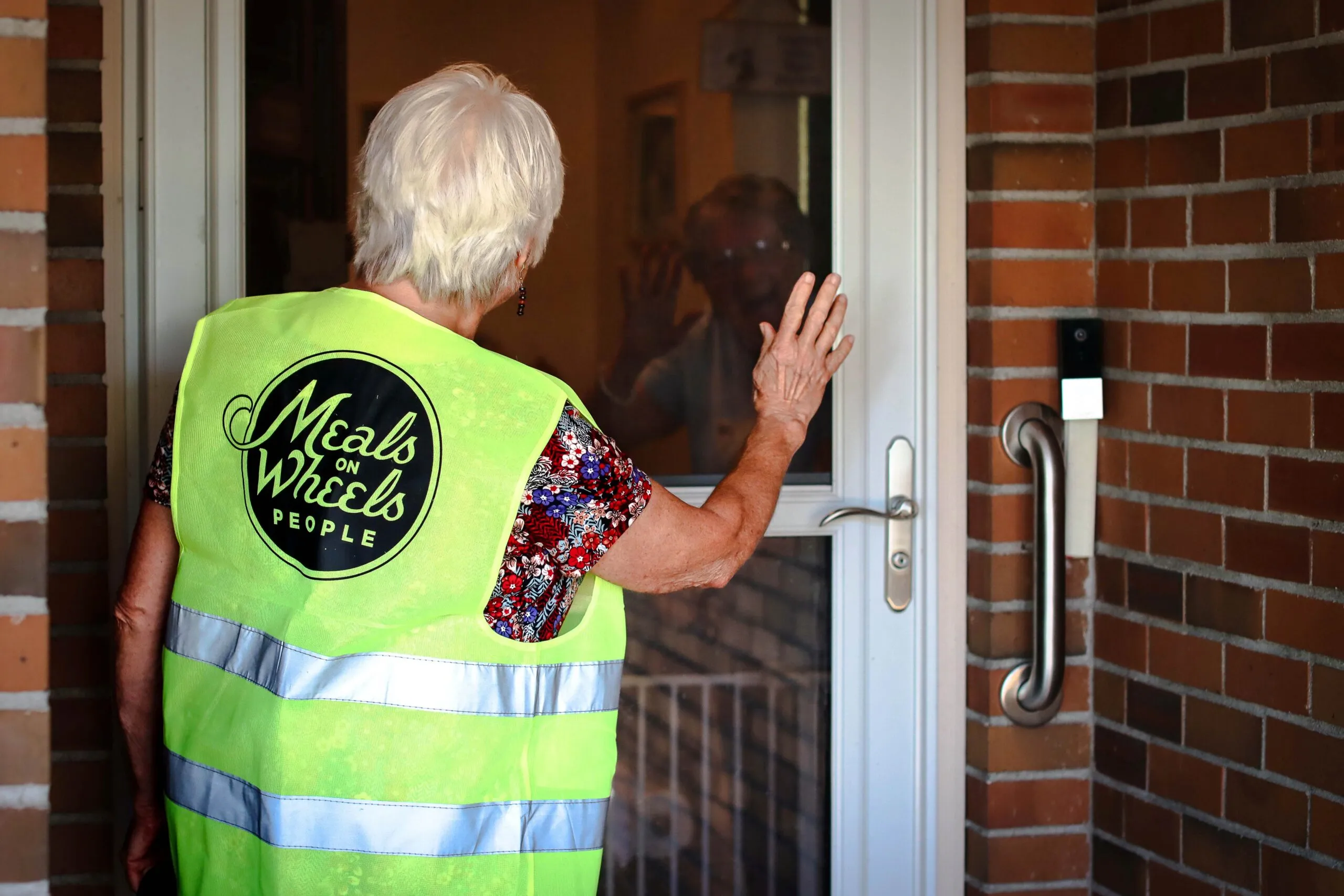 A volunteer in a bright yellow "Meals on Wheels People" vest knocks on a brick house door, greeted warmly by a resident inside. The scene conveys care and community support.