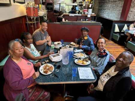 A group of six people smiling and seated around a teal diner table with plates of food. The setting is a cozy, retro-style restaurant with wood floors.