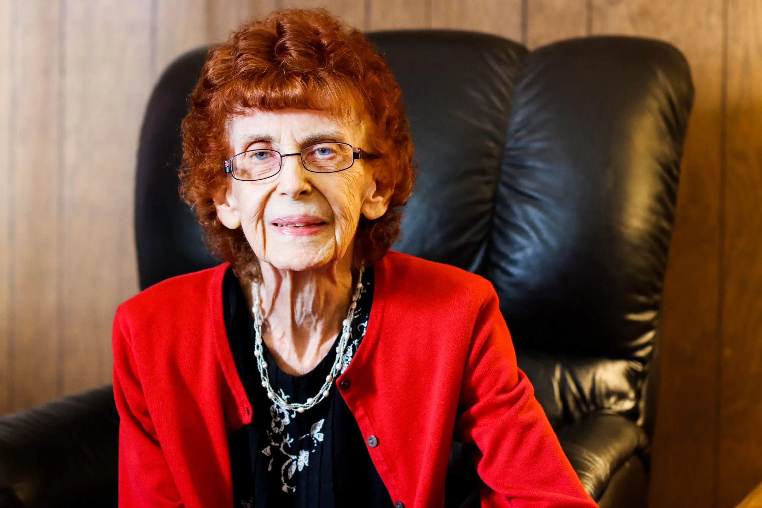 Older woman with glasses and curly red hair, wearing a red cardigan, sits on a black leather chair, smiling softly against a wooden wall background.