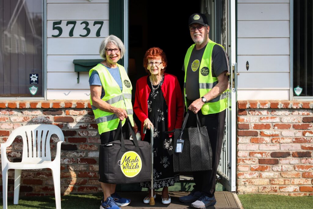 Three smiling people stand at a doorway; two in neon vests labeled "Meals on Wheels People" and an older woman in a red cardigan using a cane.