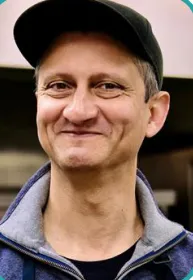 Pankaj Ramesh, wearing a black cap and gray jacket, stands in front of a blurred background. His expression conveys warmth and friendliness.