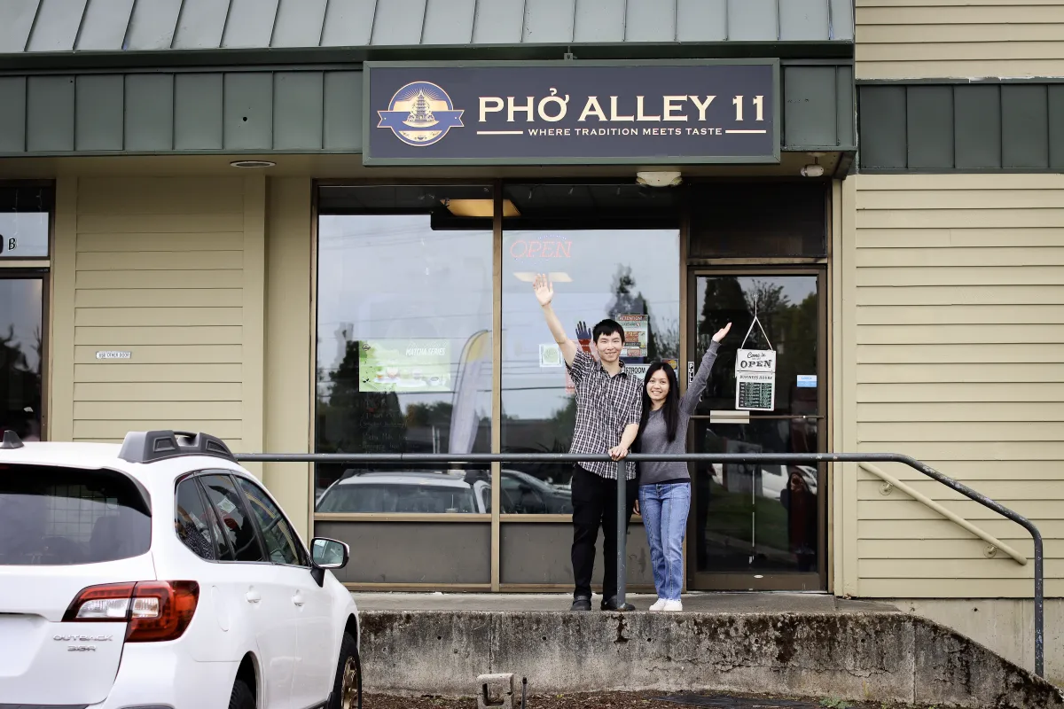 A smiling man and woman wave enthusiastically outside "Pho Alley 11," a Vietnamese restaurant. The tone is welcoming and friendly.