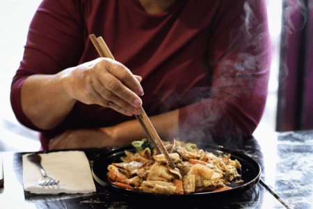 A person in a maroon shirt uses chopsticks to eat steaming stir-fry noodles with vegetables on a black plate. A fork and napkin rest on the table.