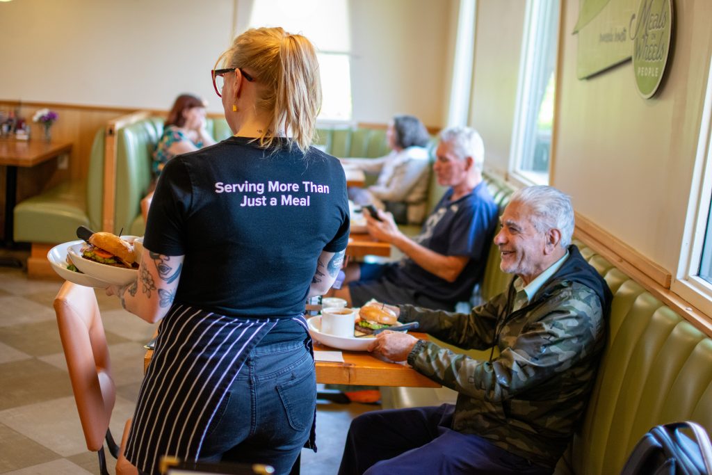 A server in a black t-shirt that reads "Serving More Than Just a Meal" carries a plate of food while an older man in a camo jacket sits at a table smiling. Other diners are visible in the background.