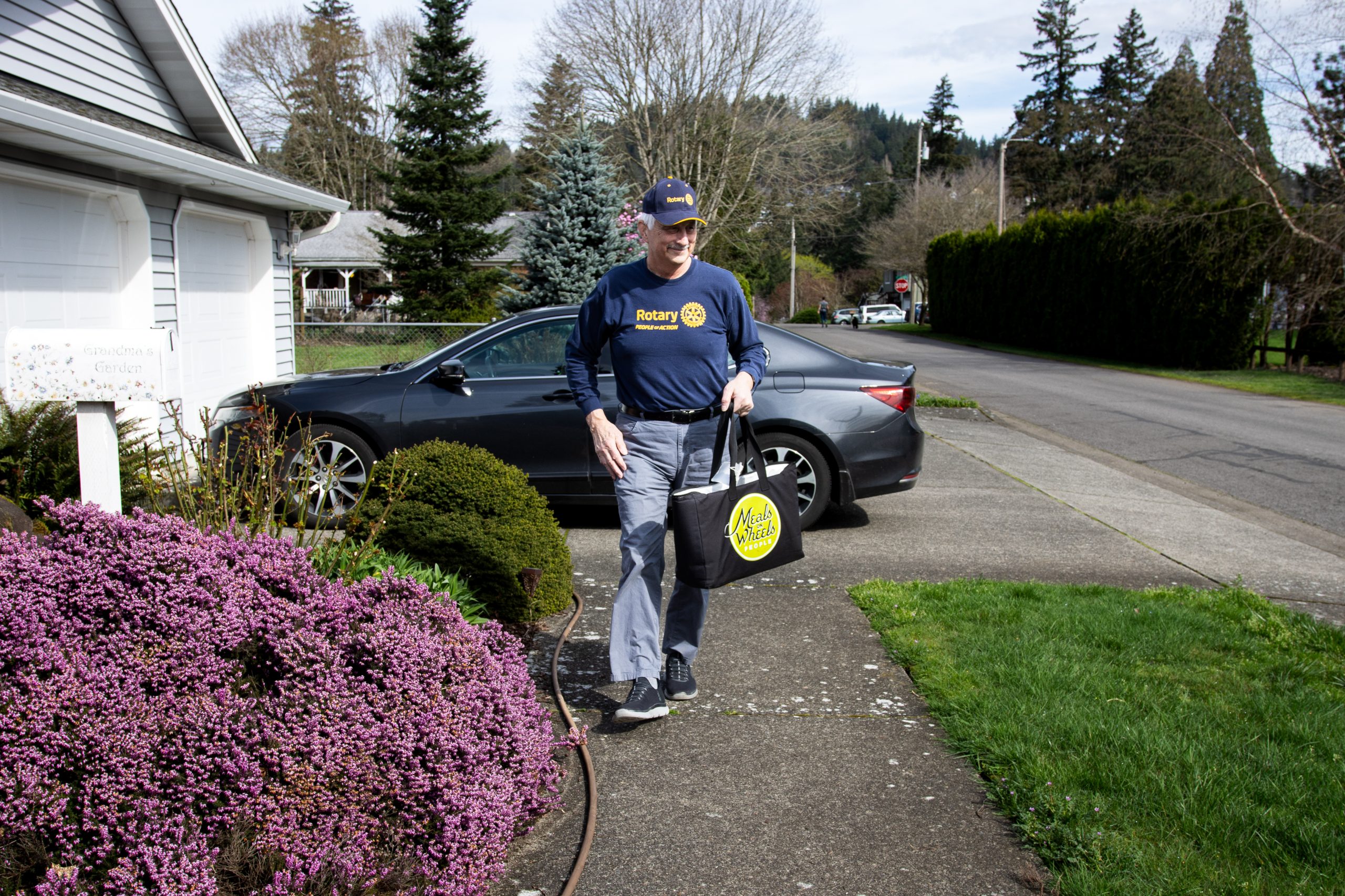 A man wearing a Rotary Club shirt and cap walks on a sidewalk near a house and a car, with landscaping featuring purple flowers nearby.