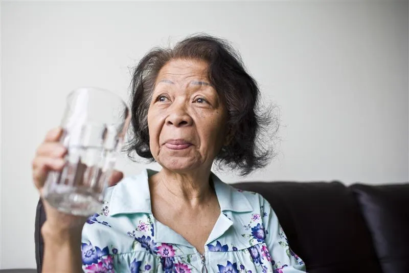 Homebound older adult with short dark hair, wearing a floral blouse, sits on a dark couch and holds up a clear glass of water with a thoughtful expression — staying hydrated during extreme heat.