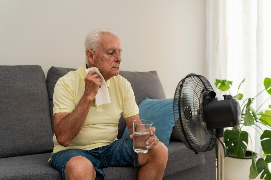 Older man refreshing himself with a glass of water and an electric fan, trying to cope with high temperature at home during summer heatwave