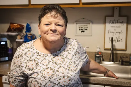 A woman with short hair stands in a kitchen, smiling and leaning against a counter.